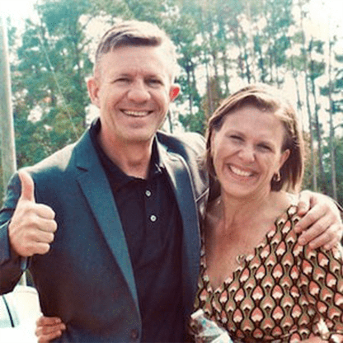 Two smiling individuals pose together outdoors, with one giving a thumbs-up