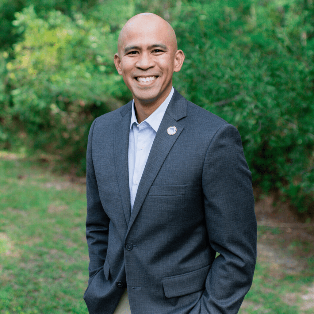 Smiling man in a gray suit stands outdoors, surrounded by greenery