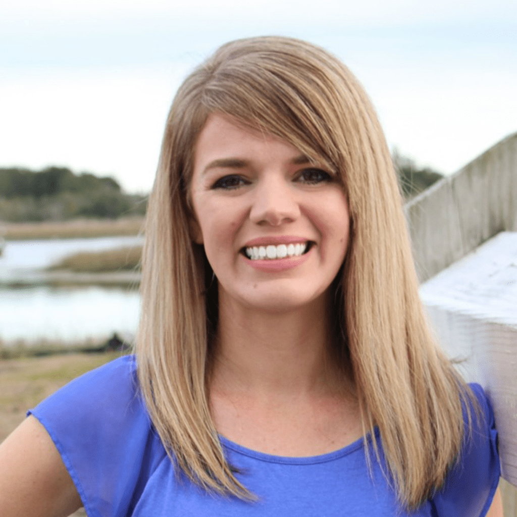 Smiling woman with long blonde hair wearing a blue shirt stands outdoors near a body of water