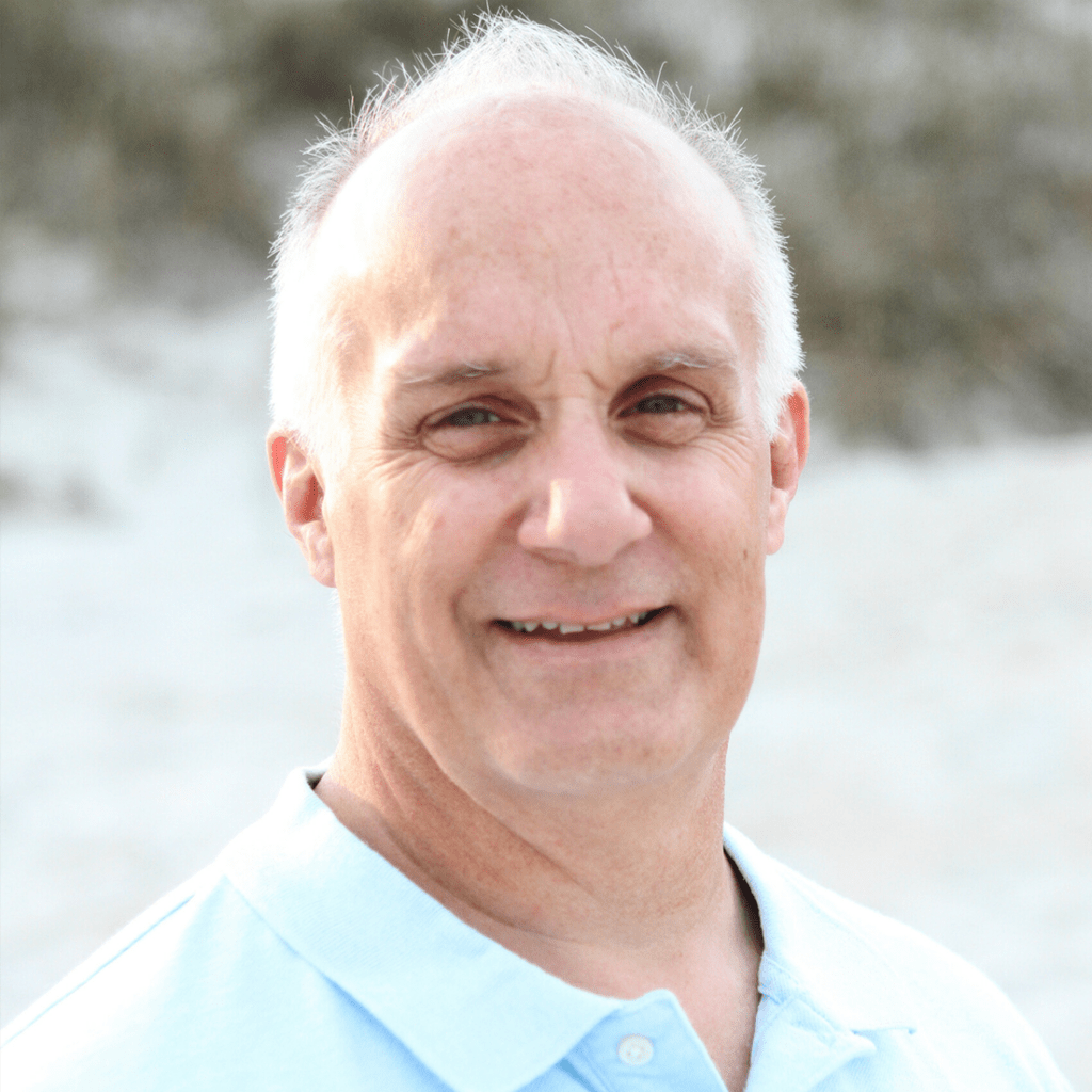 Smiling man in a light blue polo shirt stands outdoors against a sandy backdrop