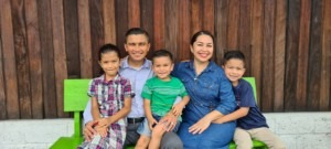 A family of five poses together on a green bench, smiling against a wooden backdrop. The image conveys warmth and togetherness.
