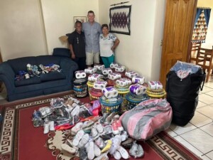 Three people stand beside a large collection of shoes and supplies in a living room, emphasizing community support and outreach efforts.