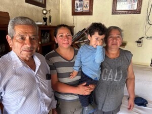 A family portrait featuring a senior man, a woman holding a young boy, and another woman, all standing together in a cozy living room setting.