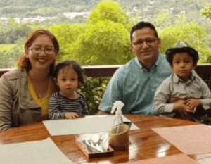 Family portrait at a restaurant table, featuring a couple with two young children against a green, scenic backdrop.