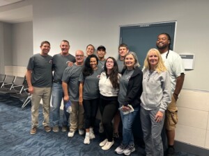 A diverse group of ten people poses together in a hallway, smiling, wearing casual gray shirts. Their cheerful demeanor suggests camaraderie and teamwork.