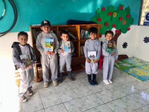 Five children in gray uniforms stand in a brightly colored classroom, holding books in front of a wooden bookshelf and a decorative tree mural.