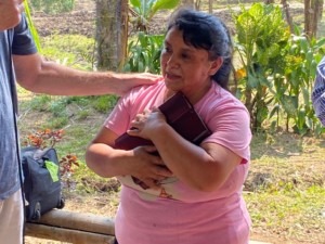 A woman in a pink shirt holds a book closely, expressing gratitude, while receiving support from a man. The setting is outdoors, surrounded by greenery.