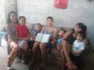 A family group sits together on a couch in a simple indoor setting, with children and adults engaged in conversation. A sense of togetherness is conveyed.