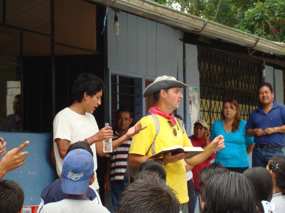 A group of people listens as a man in a yellow shirt and hat speaks