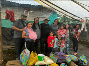 Community gathering in a makeshift shelter, featuring a diverse group of adults and children, with bags of goods displayed in front.