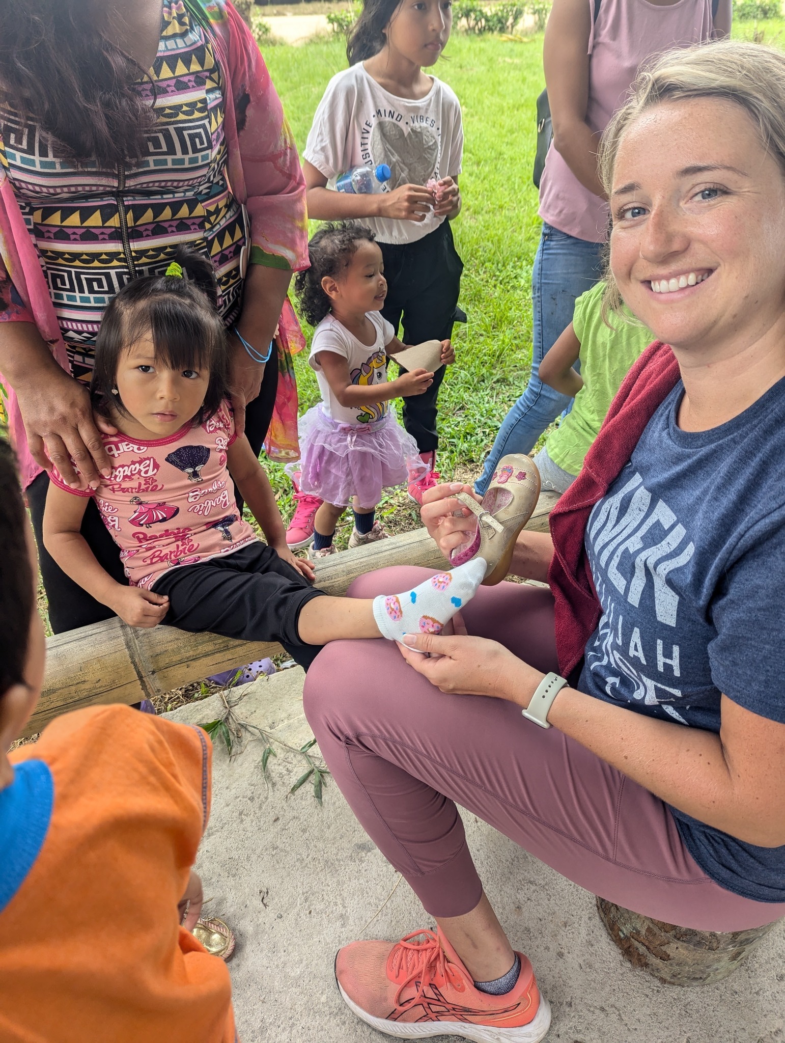 A volunteer applies a colorful bandage to a child's foot while surrounded by a group of attentive children in a grassy outdoor setting