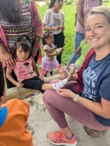 A smiling volunteer applies a bandage to a child's foot surrounded by kids and adults in a vibrant outdoor setting, highlighting community support and care.
